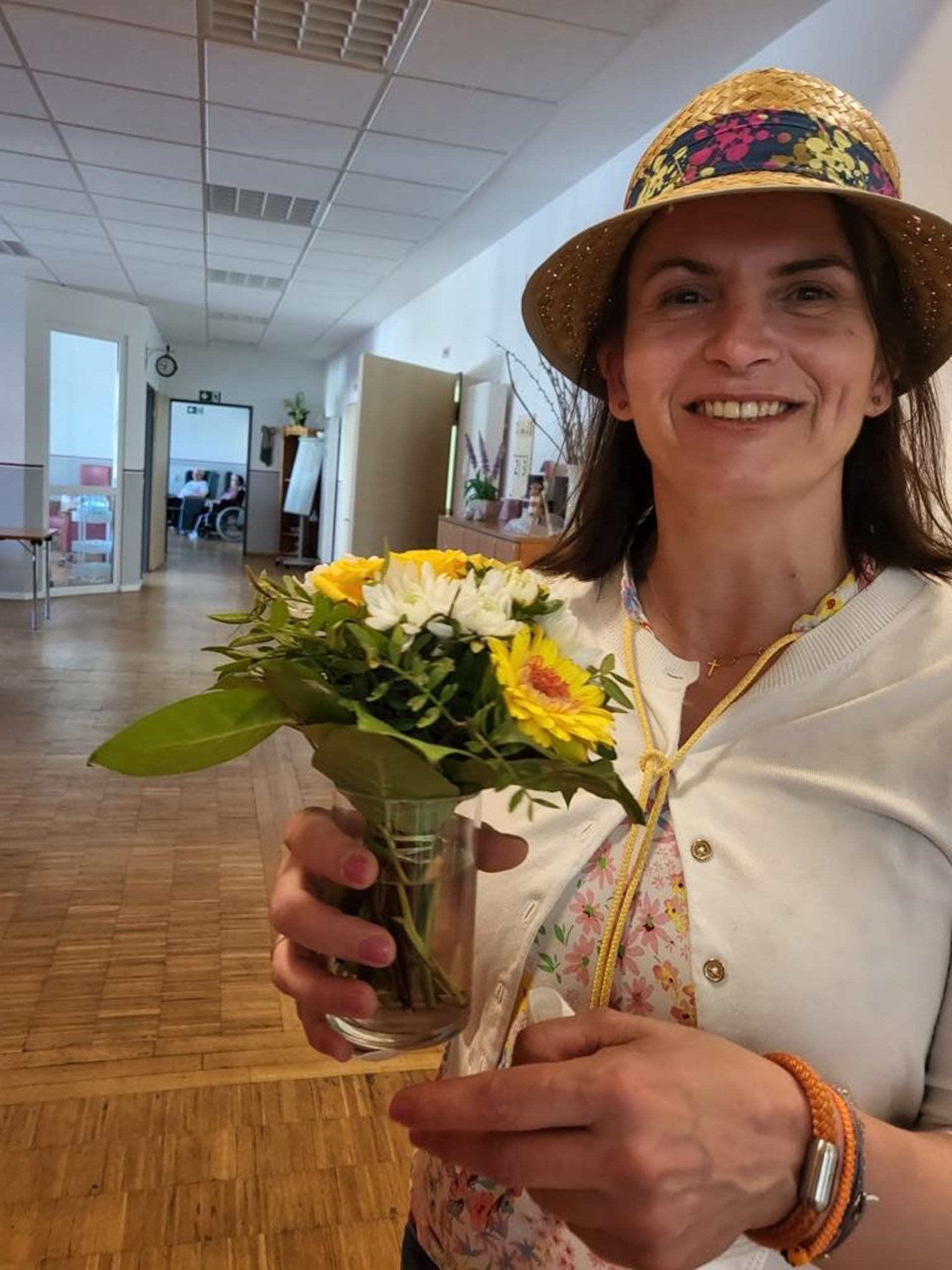 Marija Kvesiv steht in der Tagespflege, mit Hut auf, und hält lächelnd einen kleinen Blumenstrauß Richtung Kamera